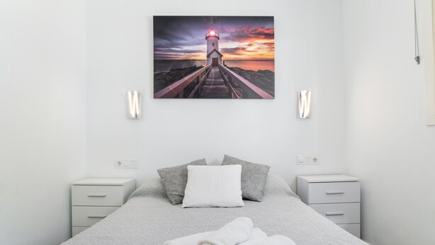 A bedroom with a bed, pillows, a nightstand, a wall-mounted picture of a lighthouse, and a power outlet.