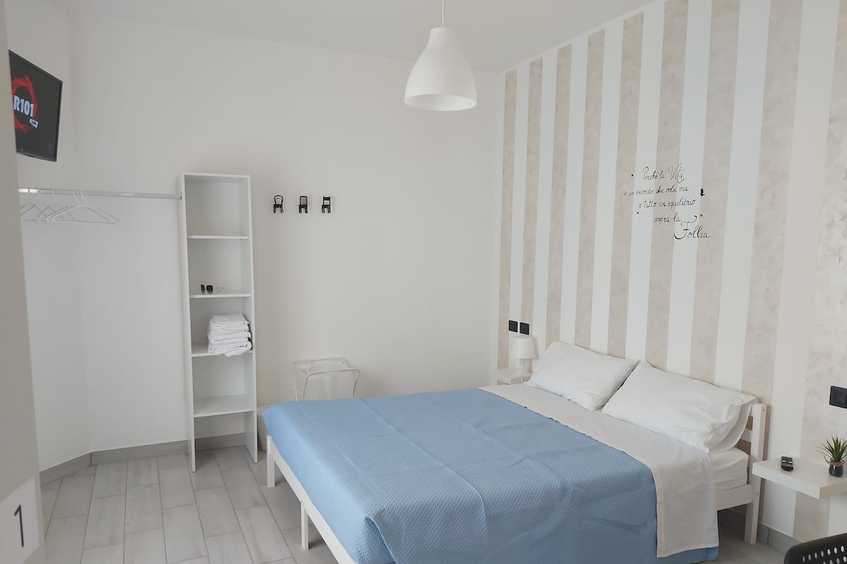 A modern hotel room with a bed, a wardrobe, a shelf with folded towels, a small table with a lamp, and a wall-mounted TV.