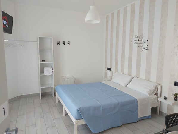 Una habitación de hotel moderna con una cama, un armario, una estantería con toallas dobladas, una mesita con una lámpara y un televisor montado en la pared.