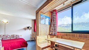 Una habitación acogedora con cama, mesa de madera y bancos, una ventana con vistas a la montaña y un estante con una radio.
