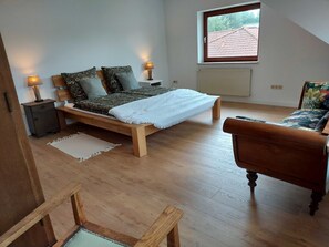 Une chambre avec un lit en bois, une table de chevet avec une lampe, une fenêtre donnant sur un bâtiment au toit rouge et une chaise en bois.