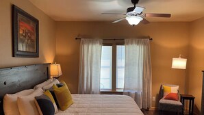 A hotel room with a bed, a ceiling fan, a window with curtains, a chair, and a lamp.