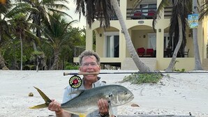 Un hombre sostiene un pez en la playa, con un edificio al fondo.