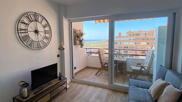 A modern living room with a large wall clock, a flat-screen TV, a sofa, and a balcony with a view of buildings and the sea. - WELCS ESTUDIO 172 EMP con piscina comunitaria (Rosas / Roses)