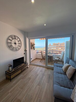 Una sala moderna con un gran reloj de pared, un televisor de pantalla plana, un sofá y un balcón con vistas a edificios y al mar.