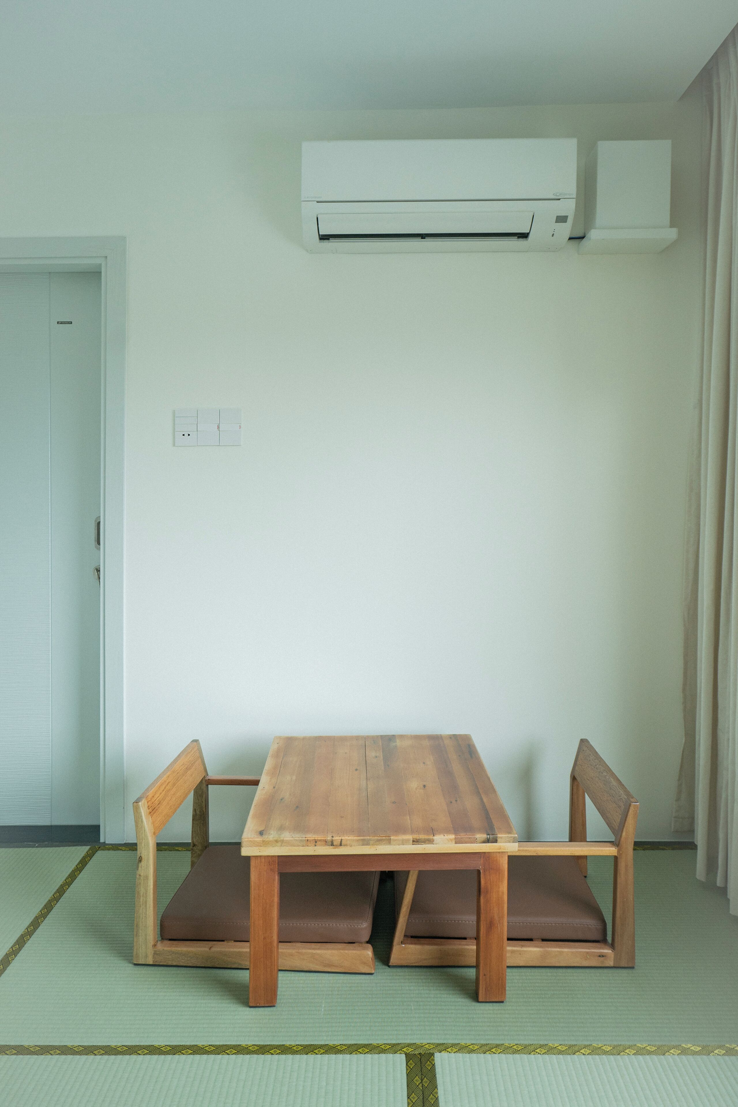 A room with a wooden dining table, two chairs, tatami mat flooring, an air conditioning unit, a door, and a light switch on the wall.