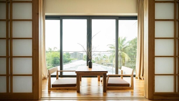 A room with a wooden table and two chairs, large windows with a view of greenery and buildings, and traditional wooden sliding doors. - Hajime Homestay (Hue)