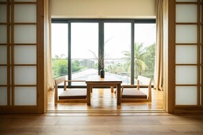 Une pièce avec une table en bois et deux chaises, de grandes fenêtres donnant sur un paysage verdoyant et des bâtiments, et des portes coulissantes traditionnelles en bois.