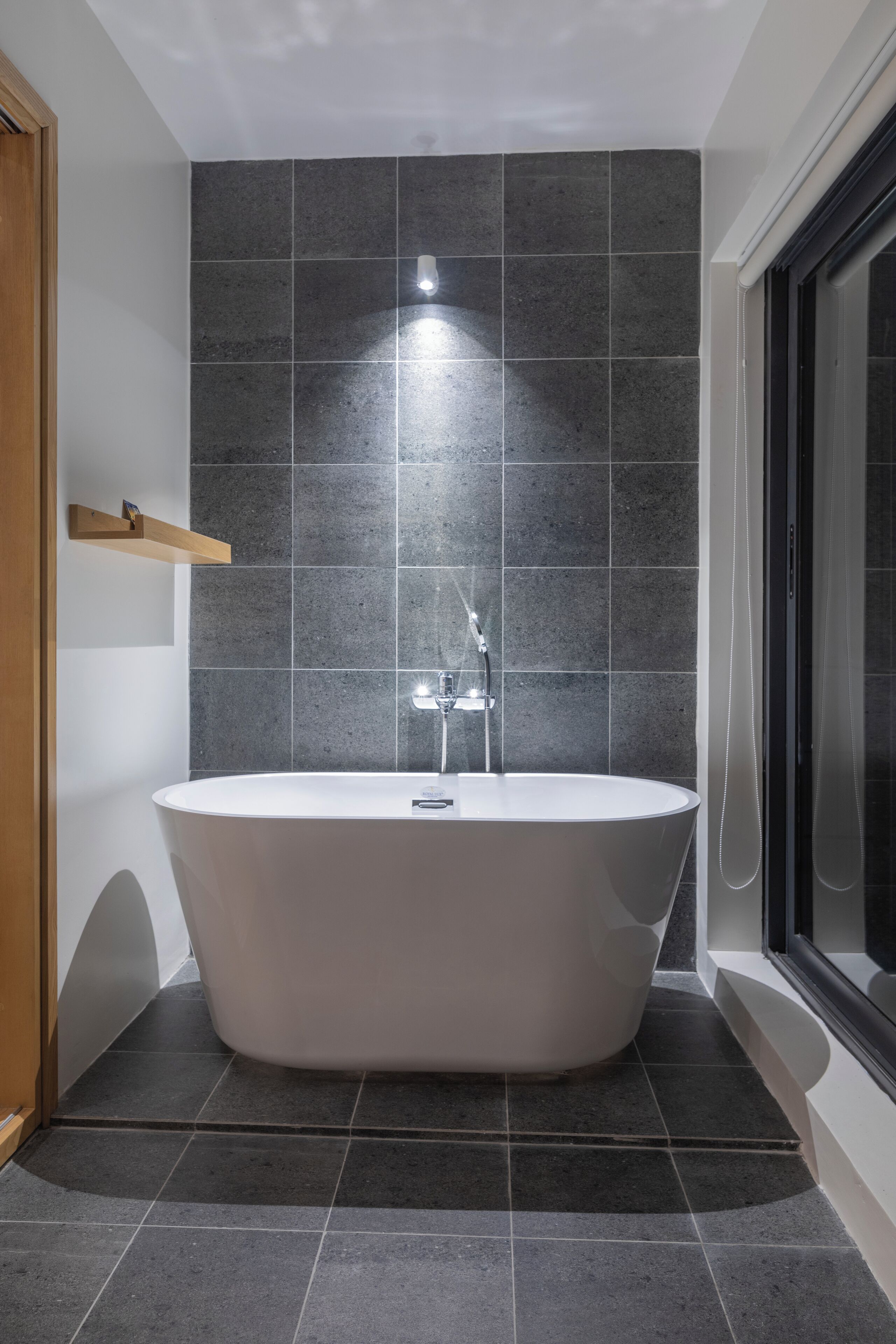 Un baño moderno con una bañera independiente, paredes de azulejos oscuros y un estante de madera.