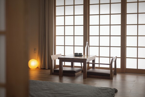 A room with a wooden table and chairs, a lit paper lantern, and a large window with a grid pattern. - Hajime Homestay (Hue)