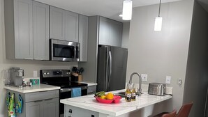 A modern kitchen with grey cabinets, a stainless steel refrigerator, a microwave, and a sink. There are two bar stools and a fruit bowl on the island. - Cozy 2-bedroom condo with WiFi, AC in-both bedrooms - Waikoloa Village (Waikoloa Village)