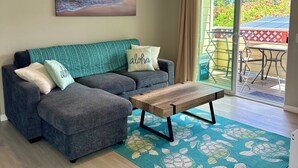 A living room with a grey sectional sofa, a wooden coffee table, a ceiling fan, a beach sunset painting, and a view of a patio with outdoor furniture. - Cozy 2-bedroom condo with WiFi, AC in-both bedrooms - Waikoloa Village (Waikoloa Village)