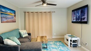 A living room with a grey sofa, a wooden coffee table, a blue rug, a ceiling fan, a TV mounted on the wall, and a beach painting. - Cozy 2-bedroom condo with WiFi, AC in-both bedrooms - Waikoloa Village (Waikoloa Village)