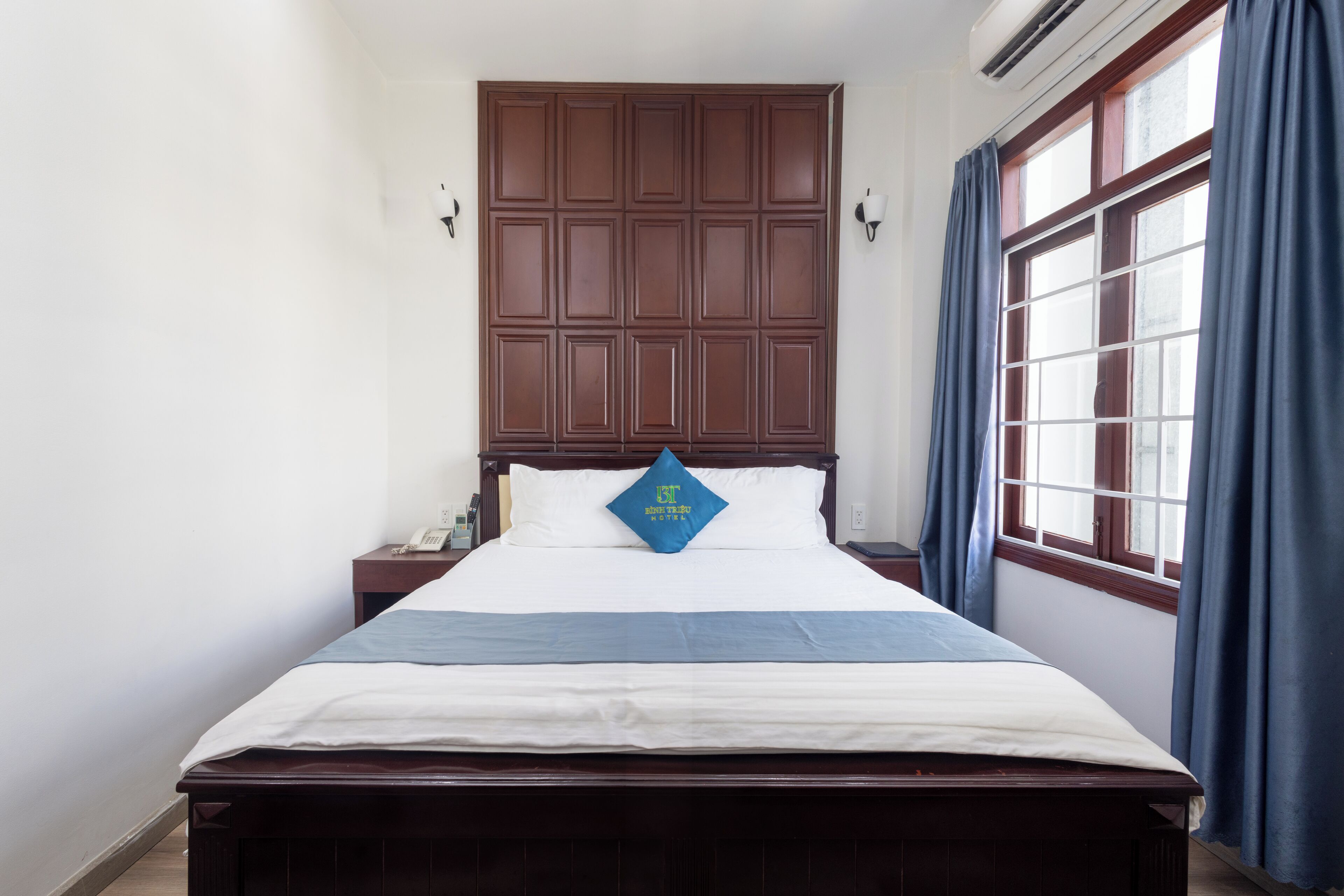 A neatly made bed with a blue pillow, a wooden headboard, a nightstand with a phone, and a window with curtains.