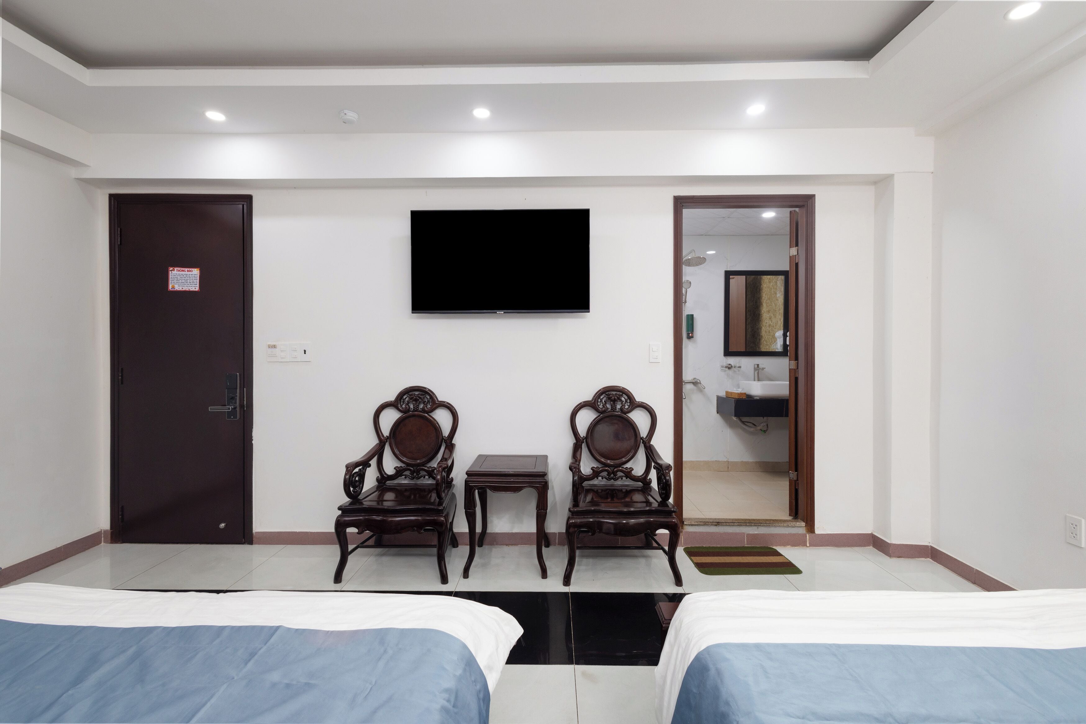 A hotel room with two beds, a flat-screen TV, two wooden chairs, a small table, and a bathroom visible through an open door.