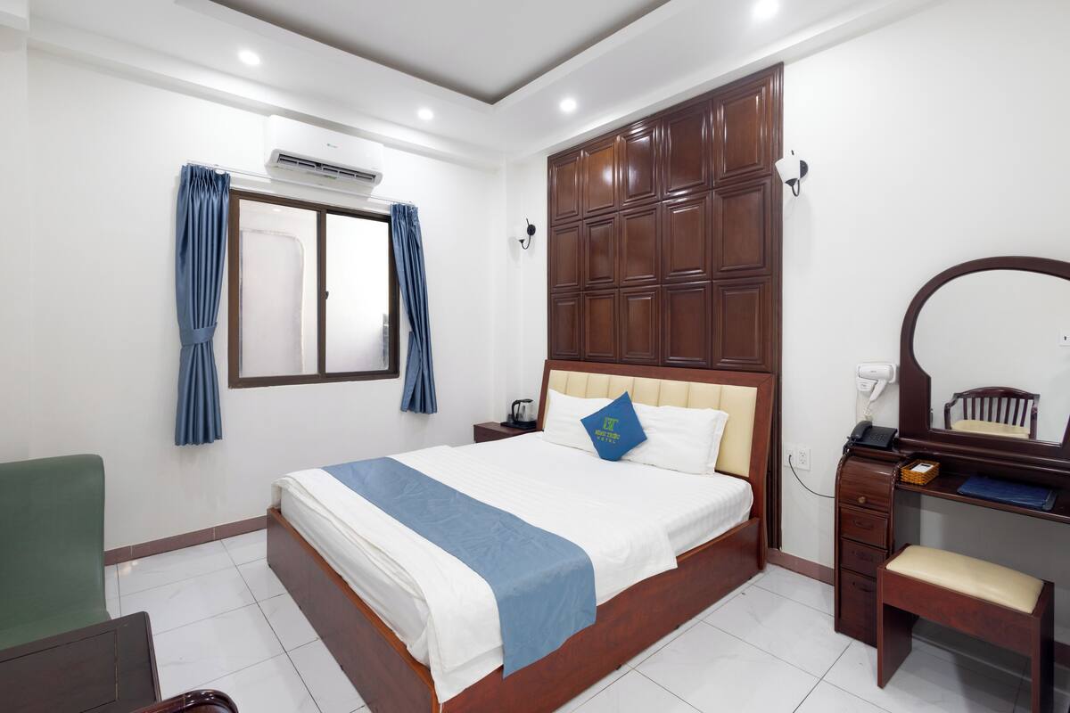 A hotel room with a large bed, a wooden wardrobe, a desk with a chair, a small table, and a mirror.