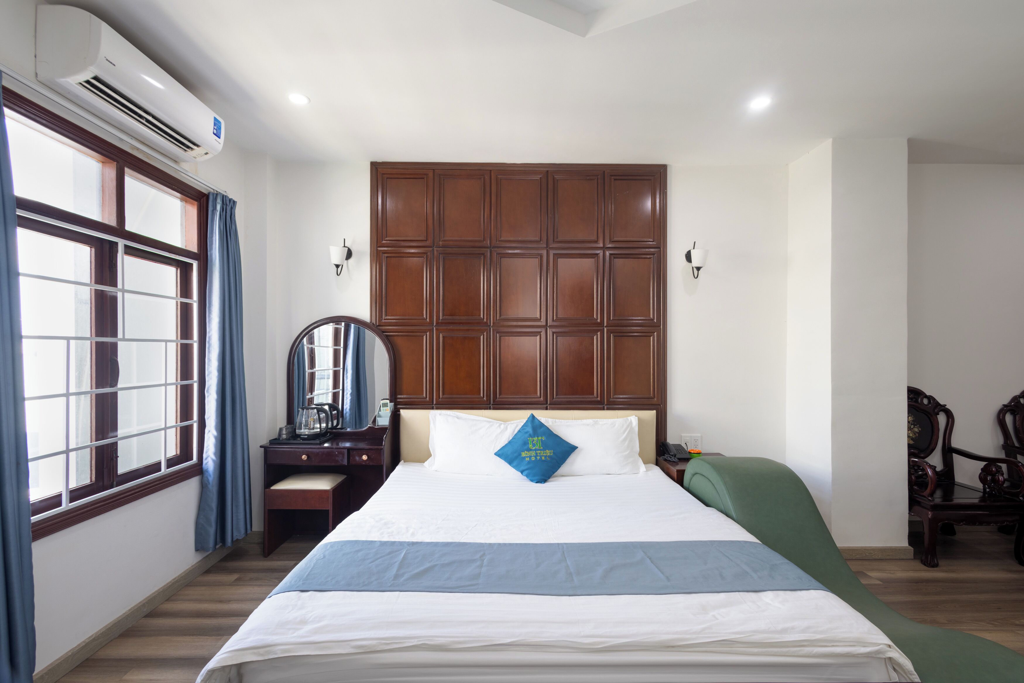 A hotel room with a large bed, a wooden headboard, a desk with a chair, a mirror, and a window with curtains.