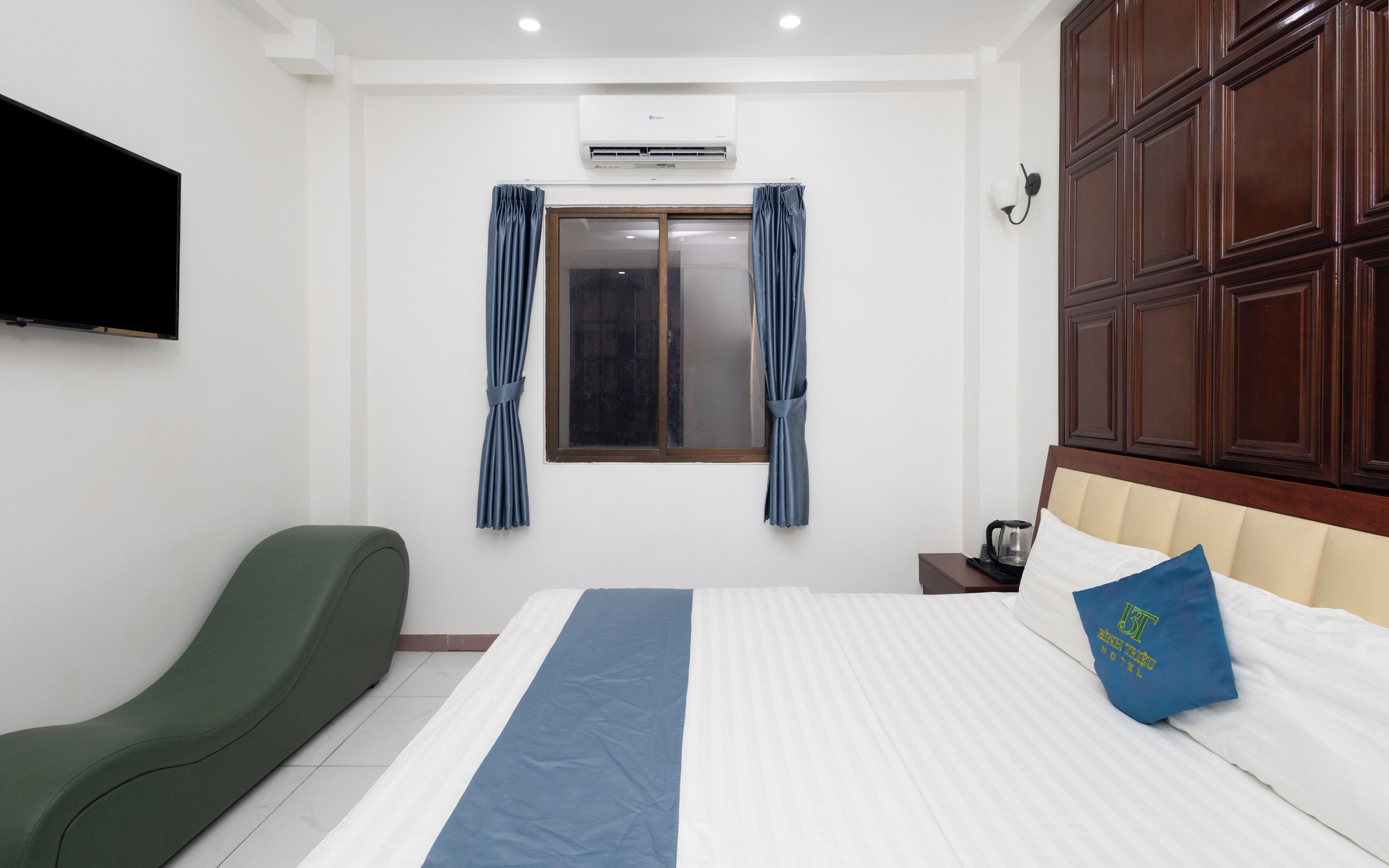 A hotel room with a bed, a green chair, a window with curtains, an air conditioning unit, and a wall-mounted TV.