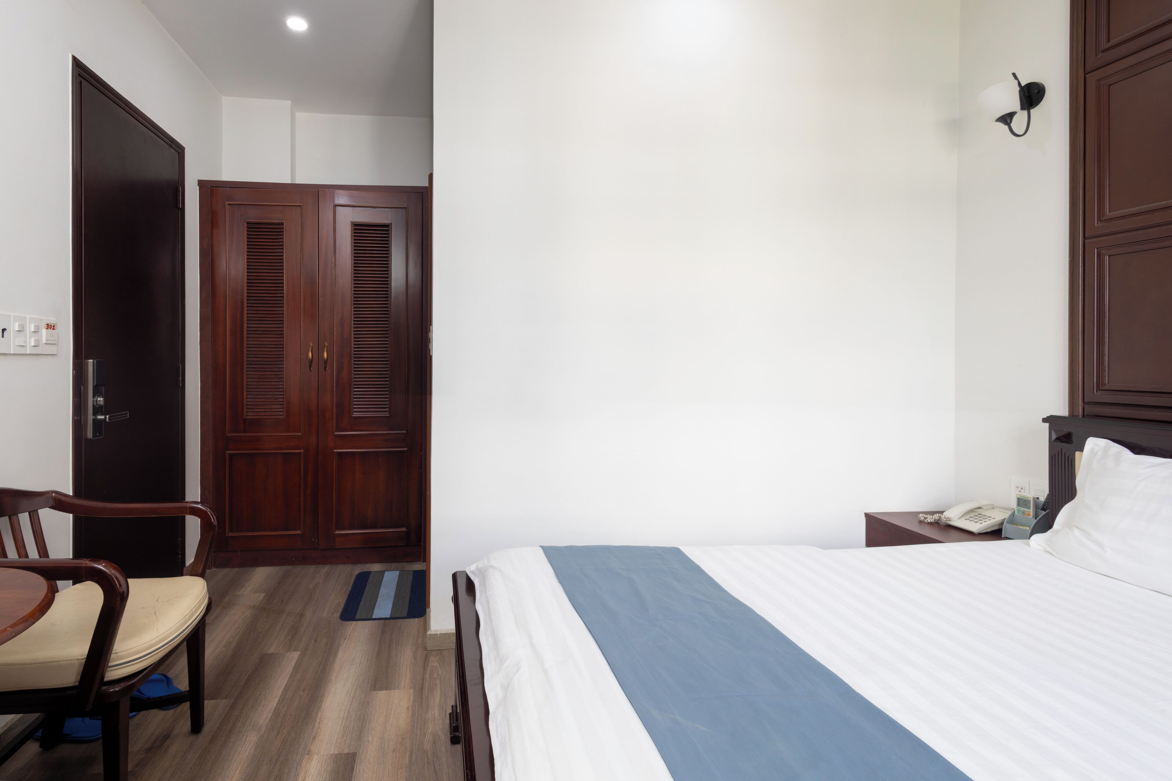 A hotel room with a bed, a desk with a chair, a wardrobe, and a bedside table with a phone.