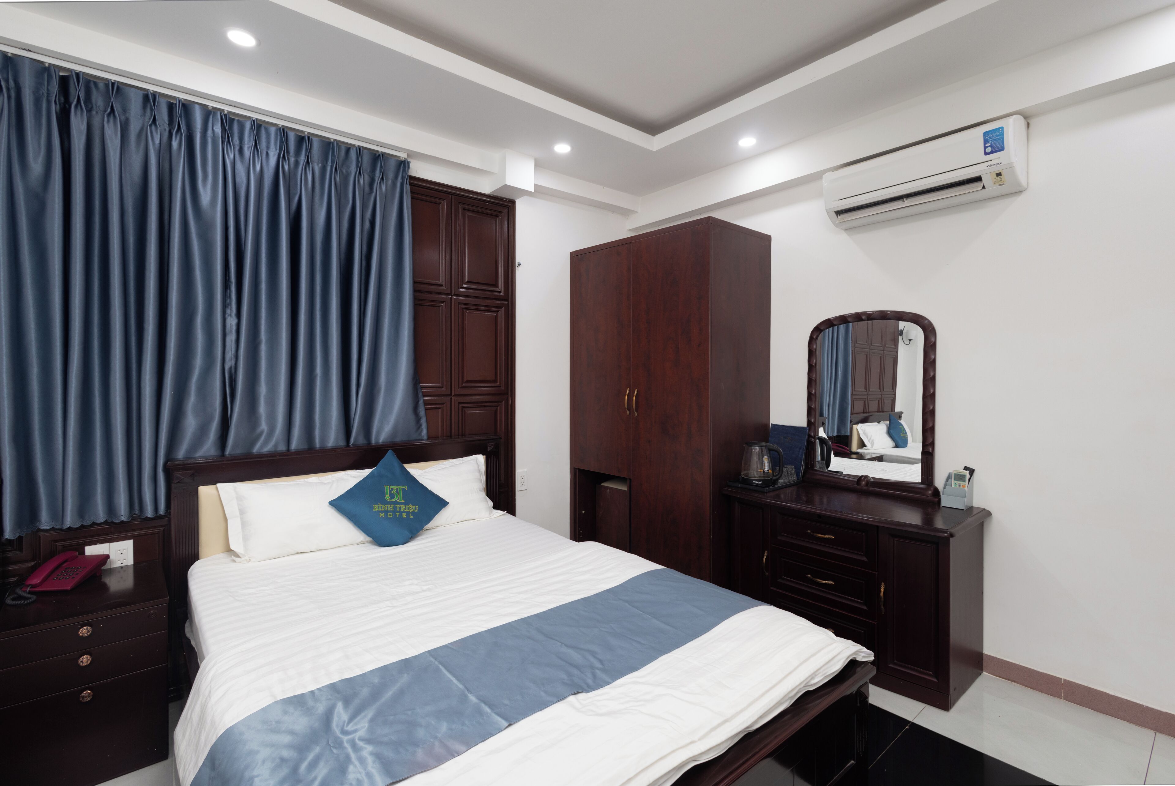 Una habitación de hotel con una cama, una mesita de noche, un armario, un espejo y un equipo de aire acondicionado.