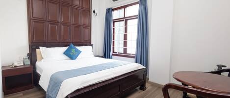 A hotel room with a wooden bed, a small bedside table, a desk, and a window with curtains.