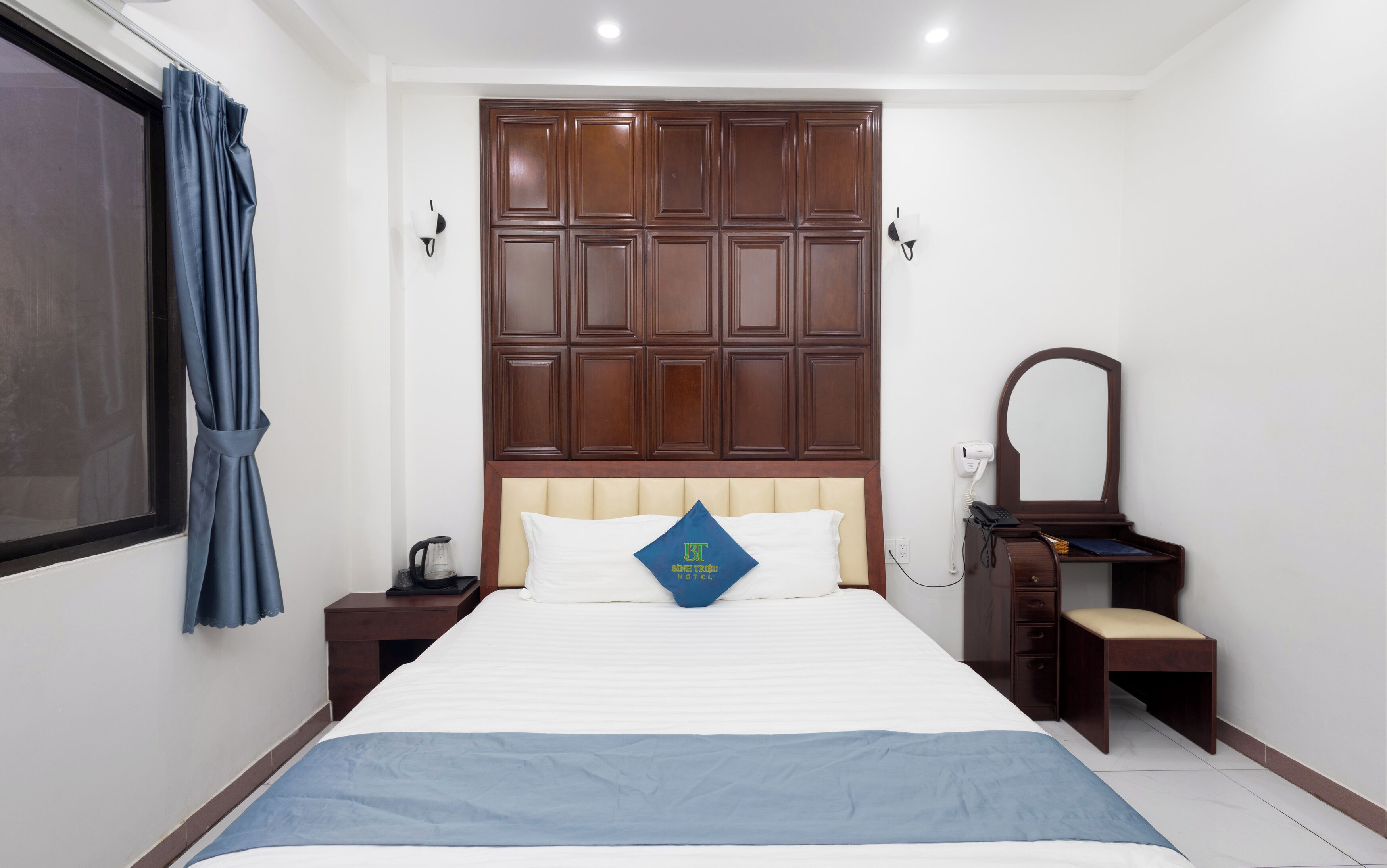 Habitación de hotel con una cama grande, cabecero de madera, mesita de noche con lámpara, un espejo y un escritorio pequeño con una silla.