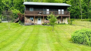 Une cabane en bois à deux étages, dotée d’un balcon, entourée d’une pelouse bien entretenue et d’une forêt dense.