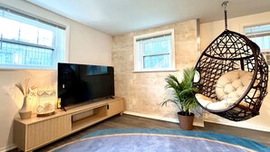 A living room with a flat-screen TV on a wooden stand, a hanging chair, a potted plant, and a large window. - Sacramento - Stylish 2BR Apt in Irving Park (Chicago)