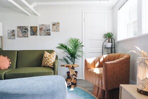 A modern living room with a green sofa, a brown armchair, and a blue ottoman. There are abstract paintings on the wall and a potted plant. - Sacramento - Stylish 2BR Apt in Irving Park (Chicago)