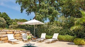 Un área junto a la piscina con un muro de piedra, dos sillas reclinables, un paraguas blanco y una piscina con baldosas.