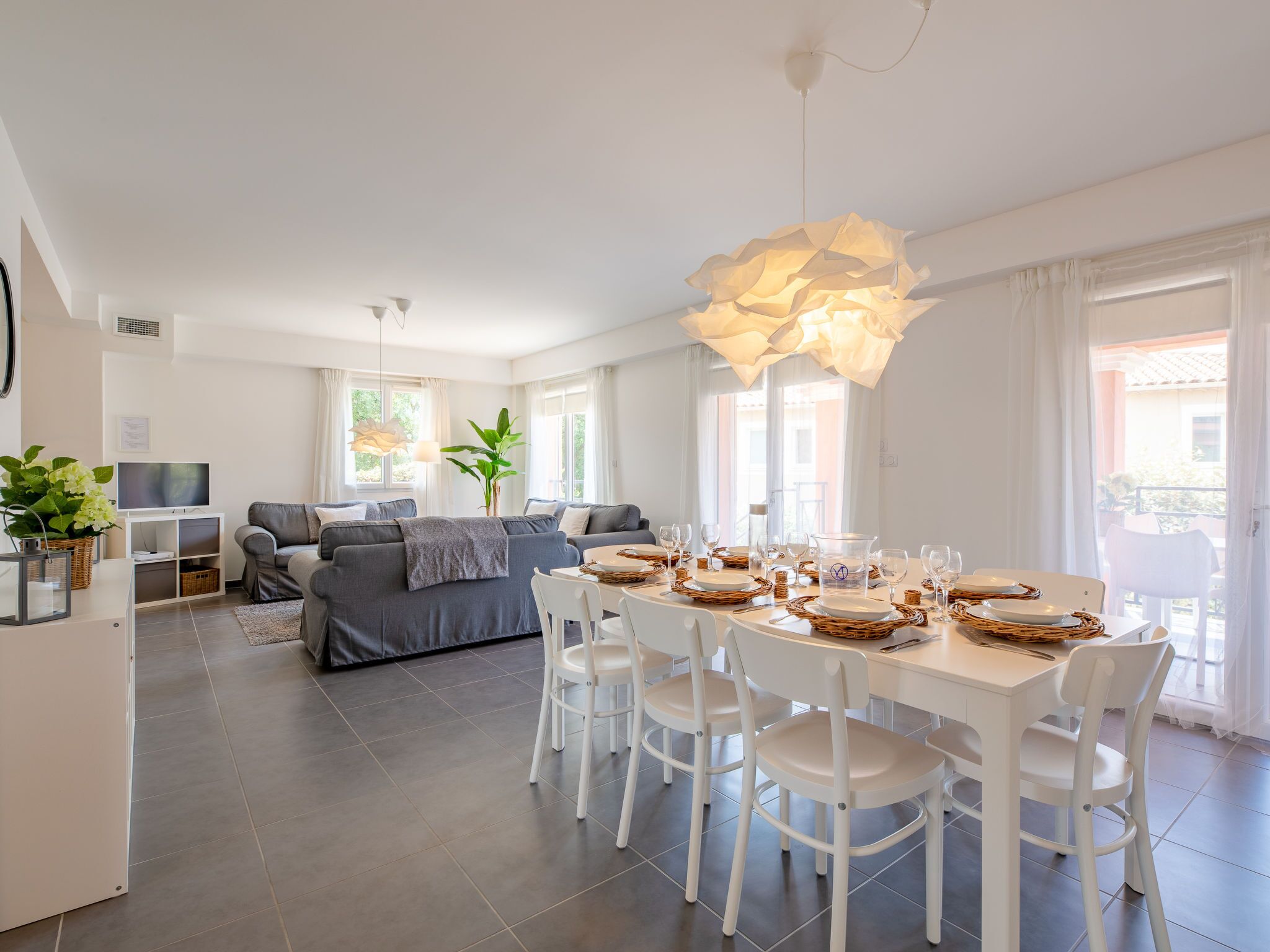 A modern, well-lit living room with a grey sofa, a dining table set for four, and a large, abstract light fixture.