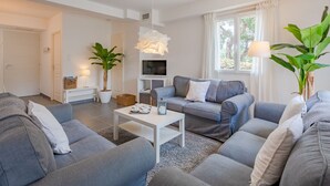 Una sala de estar moderna con sofás grises, una mesa de centro blanca y un televisor. Hay plantas en macetas y una ventana con cortinas.
