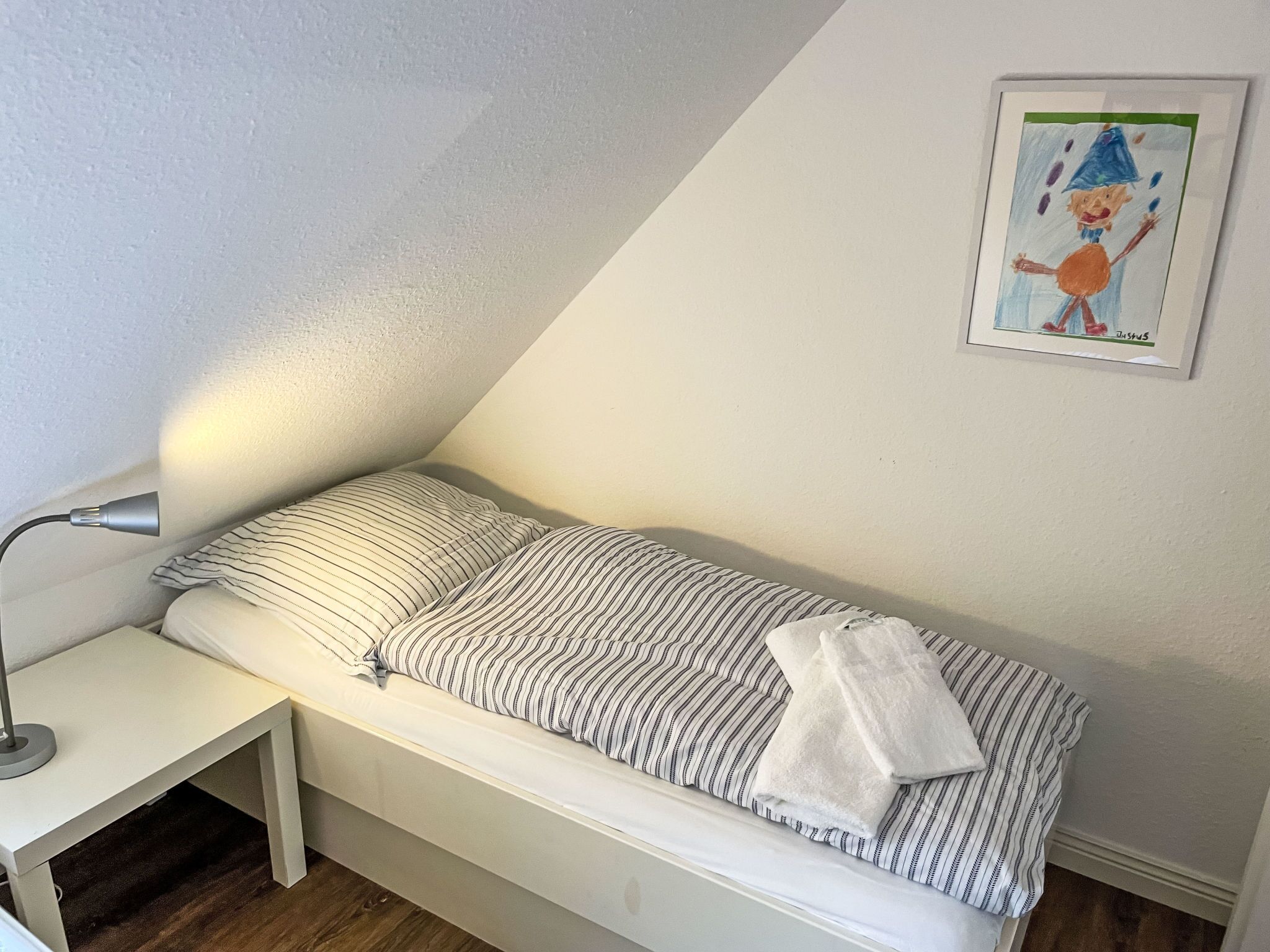 Ein kleines Zimmer mit einem Einzelbett, einer Bettdecke mit Streifenmuster, einem Nachttisch mit Lampe und einem gerahmten Bild an der Wand.