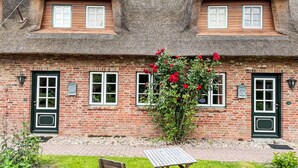 Une maison traditionnelle en briques, avec un toit de chaume, deux fenêtres avec volets et un petit jardin agrémenté d’un banc et de fleurs.