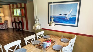 Un coin repas avec une table en bois dressée pour six personnes, des chaises blanches et un tableau représentant un voilier accroché au mur.
