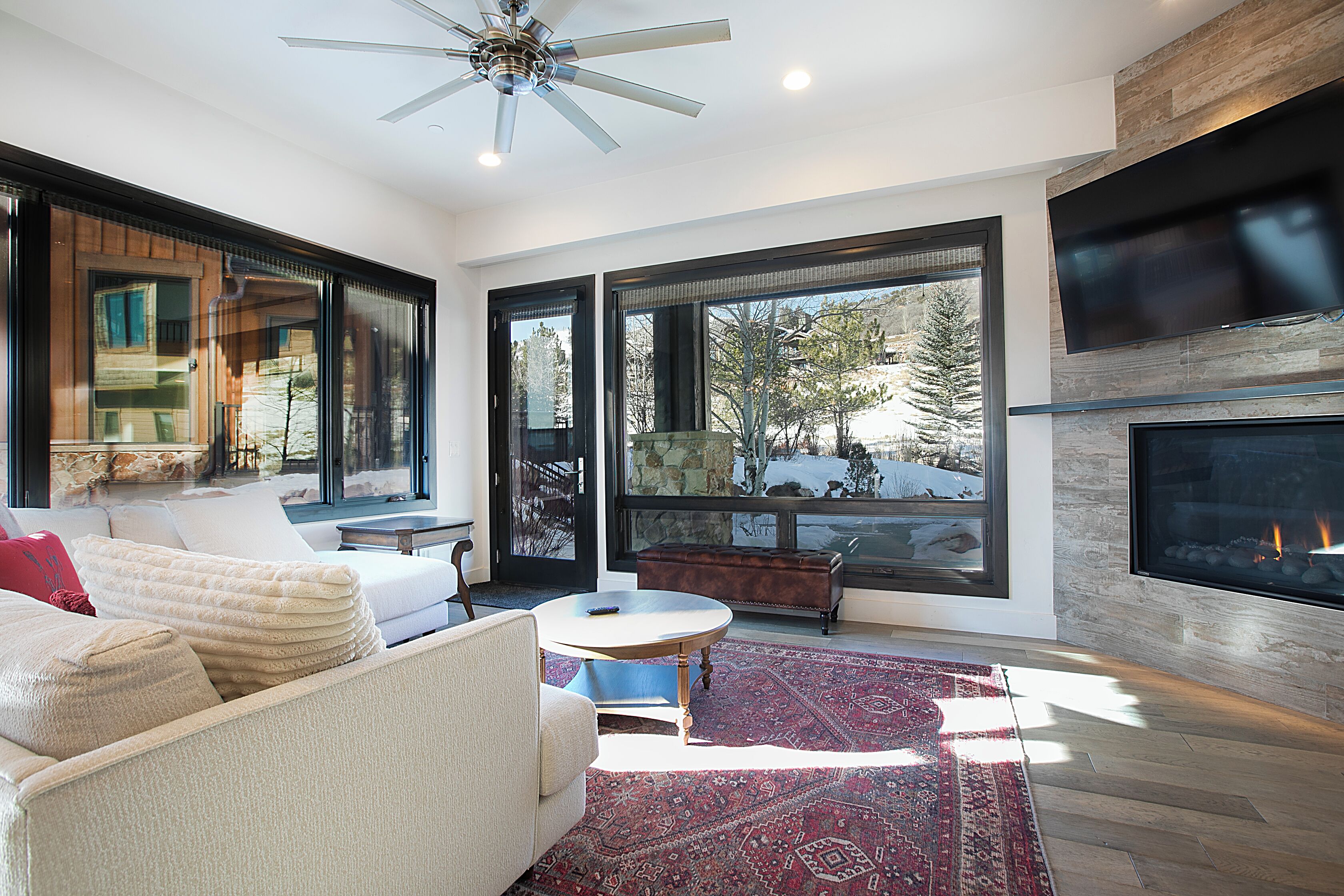 A modern living room with a fireplace, large windows, and a ceiling fan.