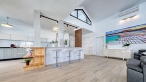 A spacious kitchen with a high ceiling, a central island with bar stools, and a large painting of a coastal scene.