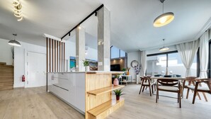 A modern kitchen with wooden shelves, a dining area with wooden chairs, and a large window with curtains.