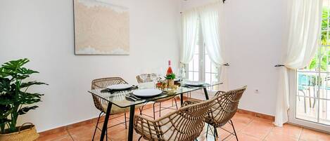 A dining area with a glass table, wicker chairs, a potted plant, and a painting on the wall.