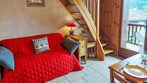 A cozy room with a red quilted sofa, a wooden staircase, a small table with a lamp, and a dining area with a table set for two. - Cosy apartment for 4 people with WIFI, TV and balcony (Saint-gervais-les-bains)