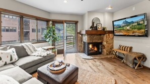 Ein modernes Wohnzimmer mit Kamin aus Stein, einem großen, an der Wand befestigten Fernseher und einer gemütlichen Sitzecke mit Sofa und Hocker.