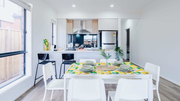 Een moderne keuken met witte kasten, een roestvrijstalen afzuigkap en een inbouwoven. Er is een eethoek met een tafel gedekt voor vier personen, witte stoelen en een tafelkleed in geel en groen.