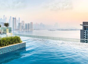 A rooftop infinity pool with a city skyline view, a glass railing, and a small planter with plants. - Relax in Comfort: Palm Studio w/ Beach Access (Dubai)