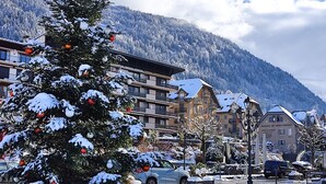 Un sapin de Noël recouvert de neige, situé sur une place de ville, avec des bâtiments et une montagne en arrière-plan.