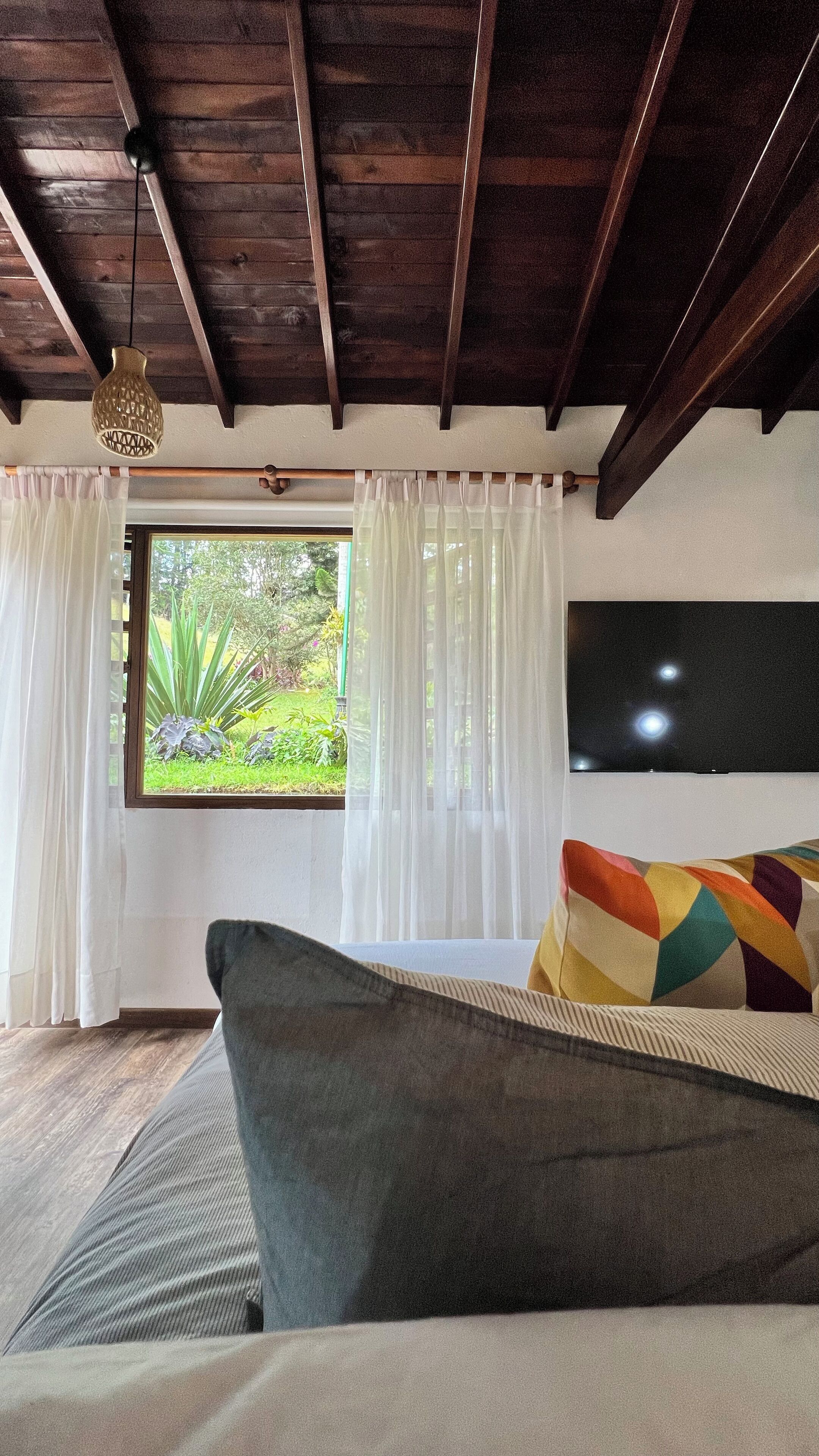 Ein Schlafzimmer mit Bett, einem Fenster mit Vorhängen, einem an der Wand befestigten Fernseher und einer Holzbalkendecke.
