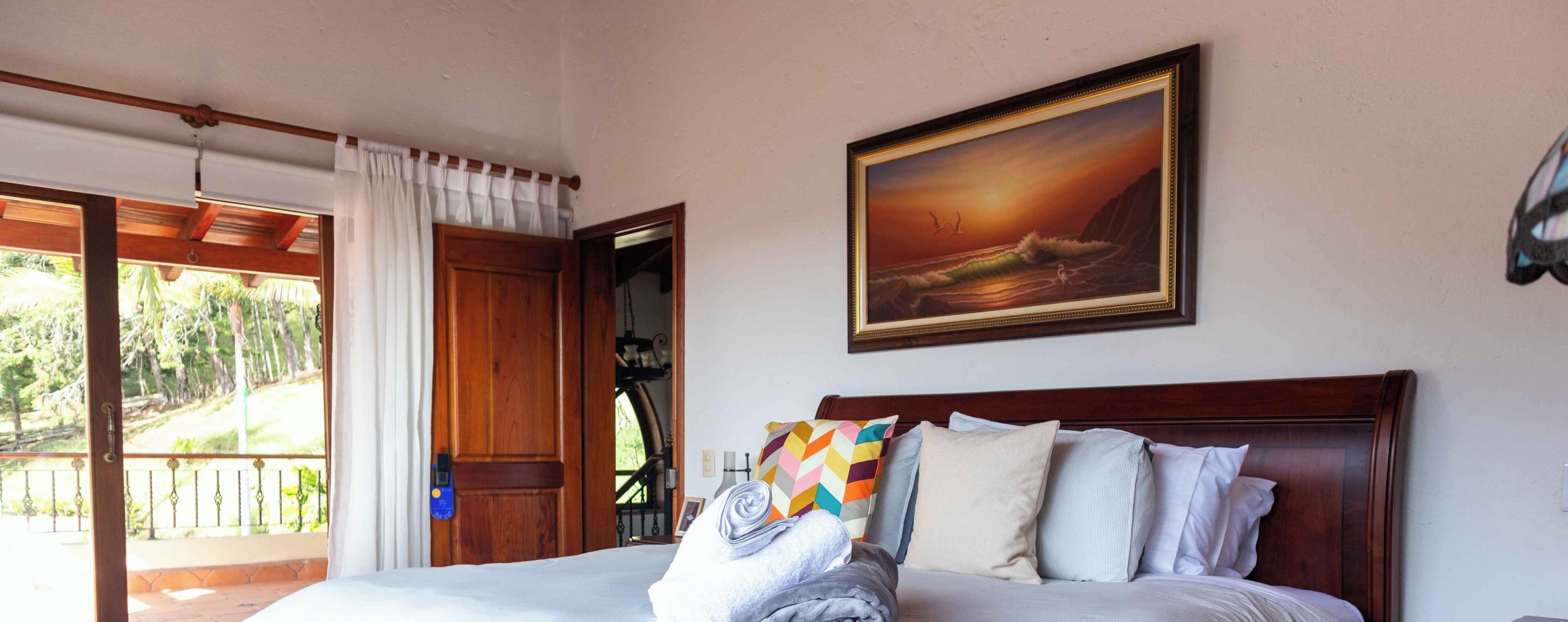 Quarto de hotel com uma cama, uma pintura de pôr do sol sobre o oceano e uma varanda com vista para o verde.