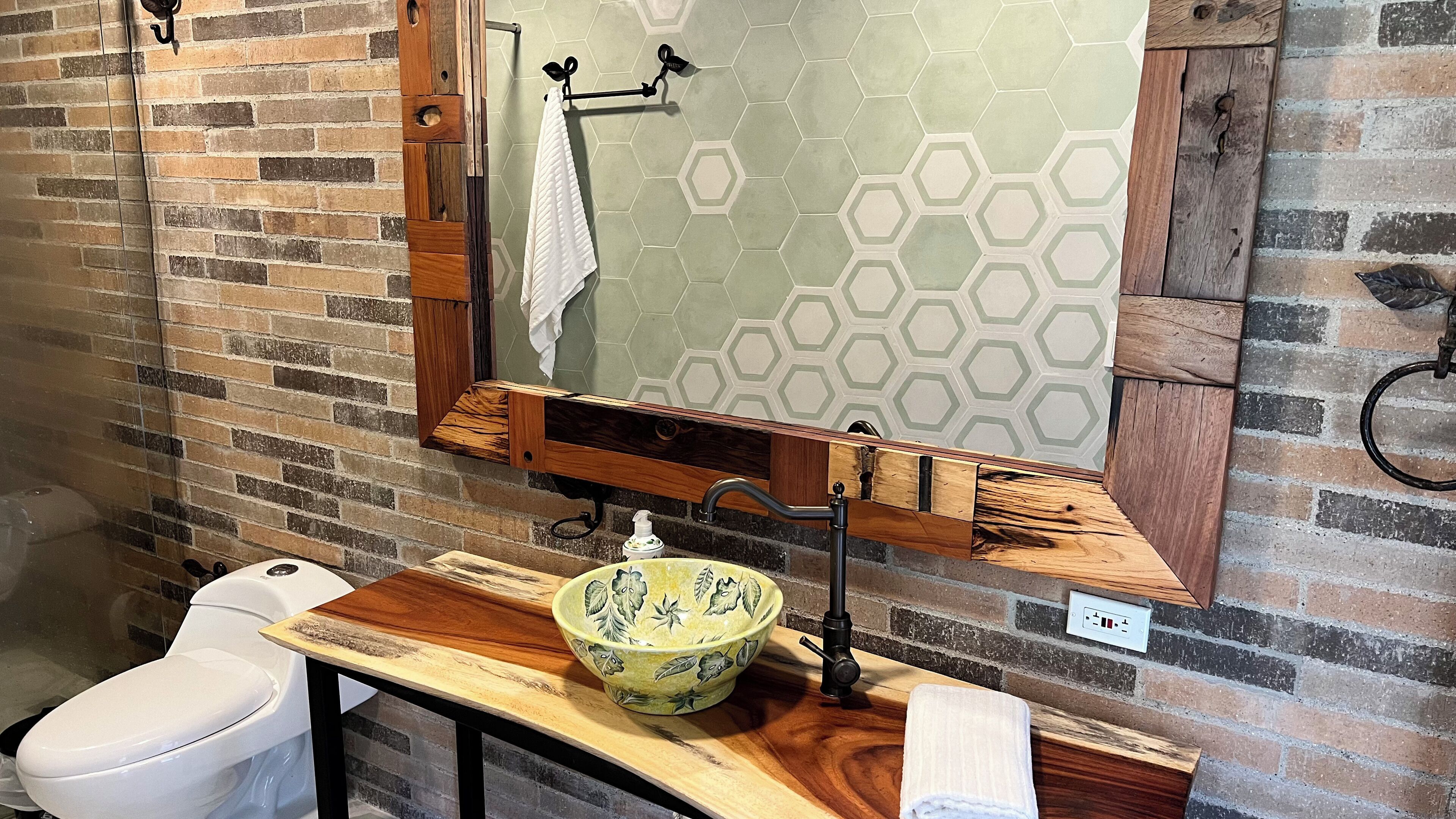 A bathroom with a wooden vanity, a green ceramic bowl, a white toilet, and a mirror with a hexagonal tile backsplash.