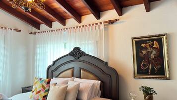 A bedroom with a wooden ceiling, a bed with white bedding and colorful pillows, a nightstand with a vase and a glass, and a framed painting on the wall.
