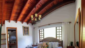 Un dormitorio con techo de madera, una cama con cabecera de madera, una araña, una ventana con cortinas y un cuadro en la pared.