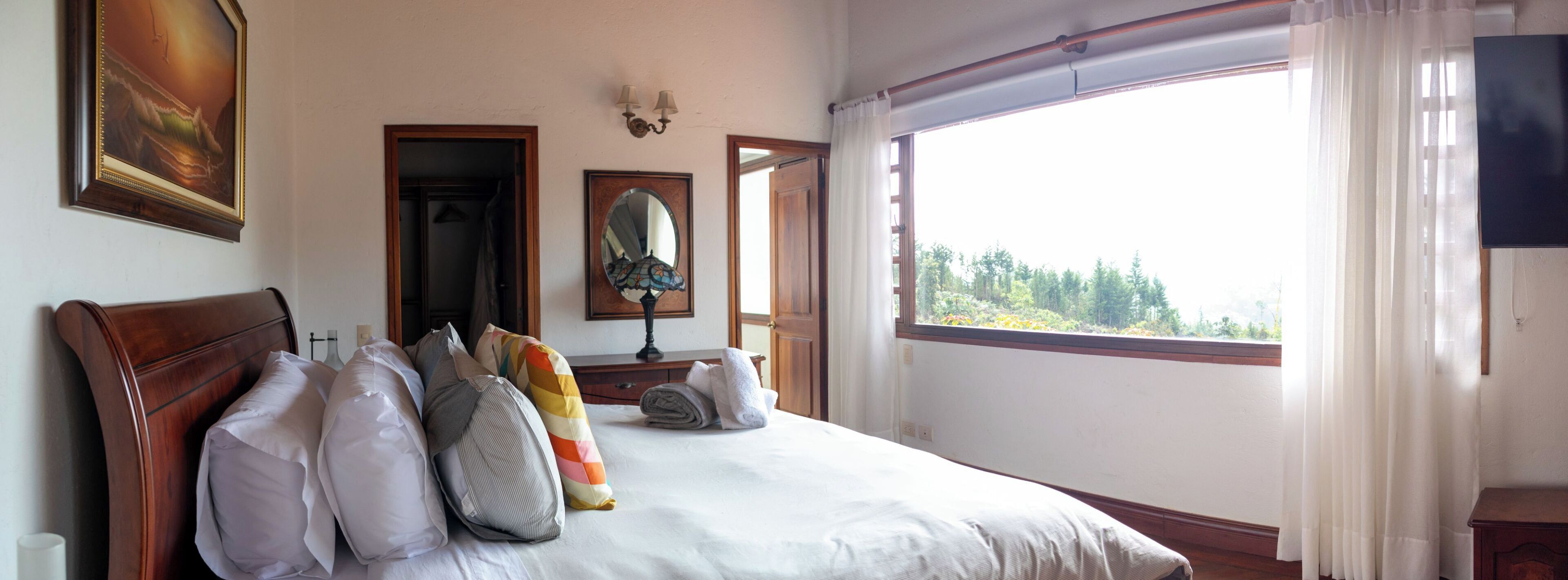 Un dormitorio con una cama grande, cabecera de madera, un espejo, una mesita de noche con lámpara y una ventana con vista a árboles.